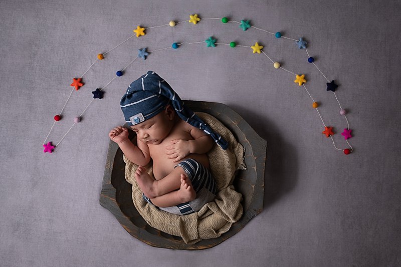 Newborn boy with blue pants and hat in a bowl with felted rainbow stars