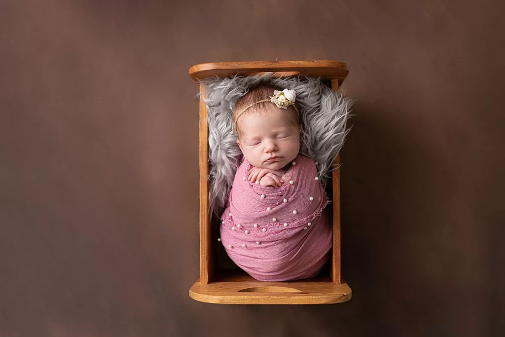 newborn girl wrapped in pink with pearl wrap in a wooden bed with gray fur