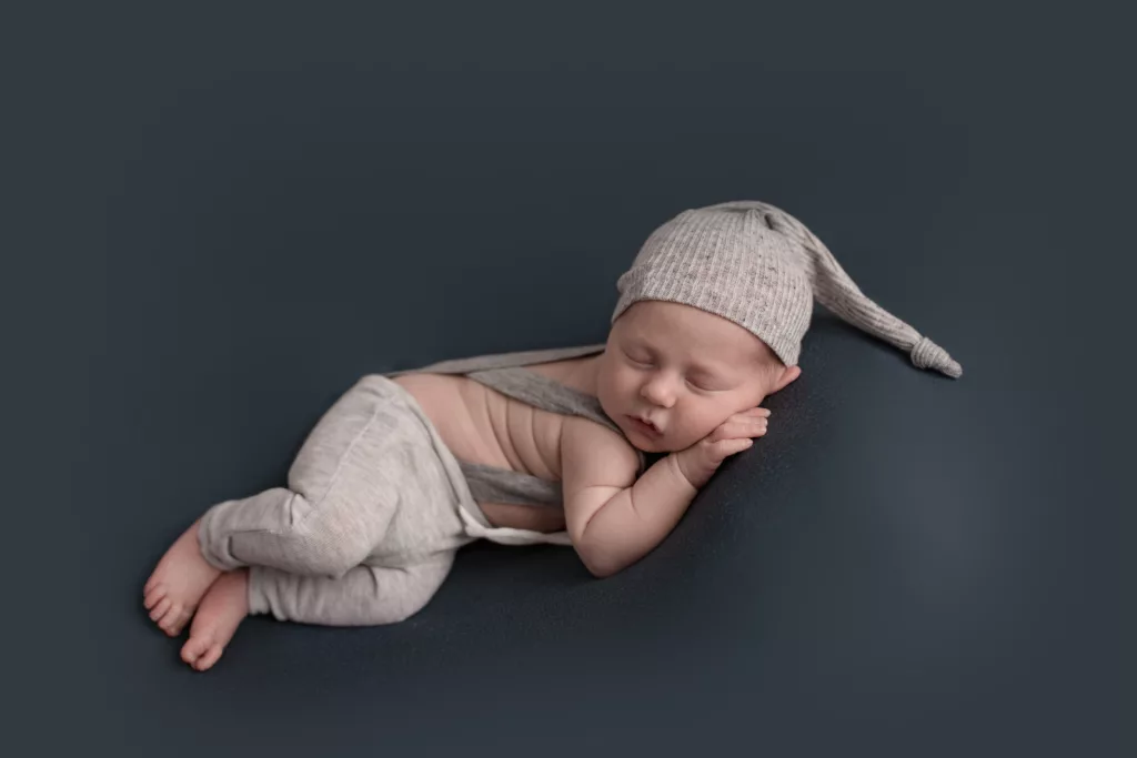 Newborn boy with a gray overalls and sleepy cap on a blue backdrop