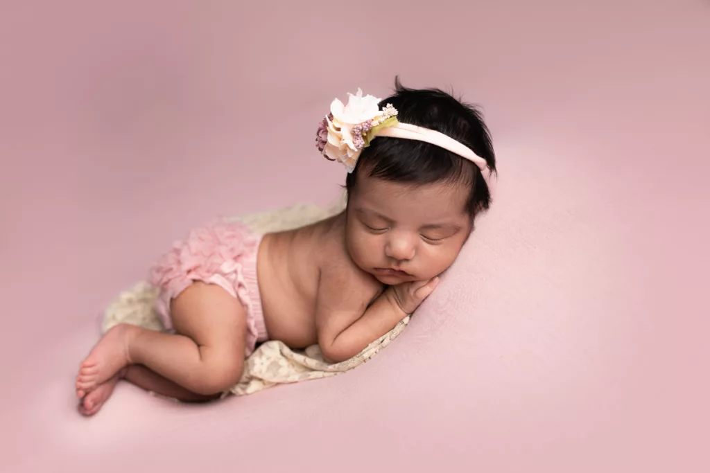 Newborn girl with a large bow and ruffled shorts asleep on a pink backdrop