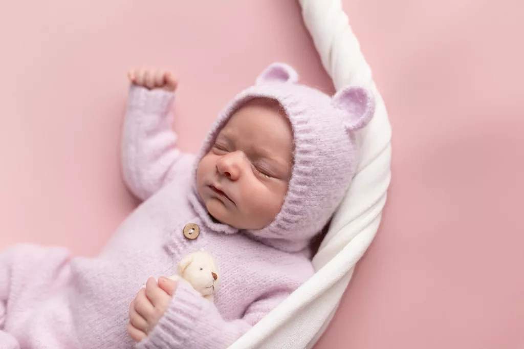 newborn girl in purple bear knit outfit on pink blanket